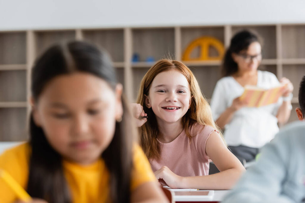 selective focus of redhead schoolgirl smiling at camera in classroom - Foto, Imagem