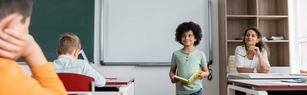african american boy standing with book near teacher and classmates, banner - Фото, изображение