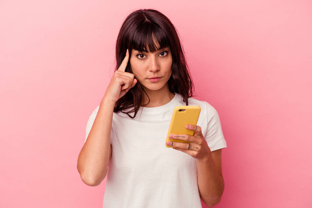 Nuori valkoihoinen nainen, jolla on vaaleanpunaiselle taustalle eristetty matkapuhelin, osoittaa sormella ohimoa ja ajattelee keskittyen tehtävään.. - Valokuva, kuva