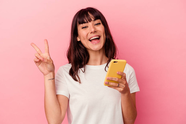 Giovane donna caucasica in possesso di un telefono cellulare isolato su sfondo rosa gioioso e spensierato che mostra un simbolo di pace con le dita. - Foto, immagini