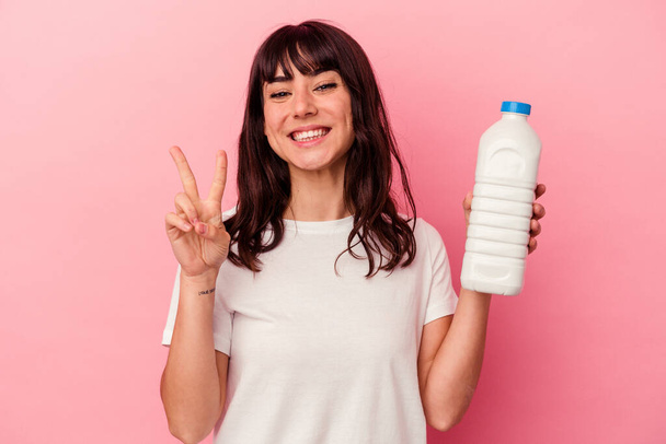 Joven mujer caucásica sosteniendo una botella de leche aislada sobre fondo rosa mostrando el número dos con los dedos. - Foto, Imagen