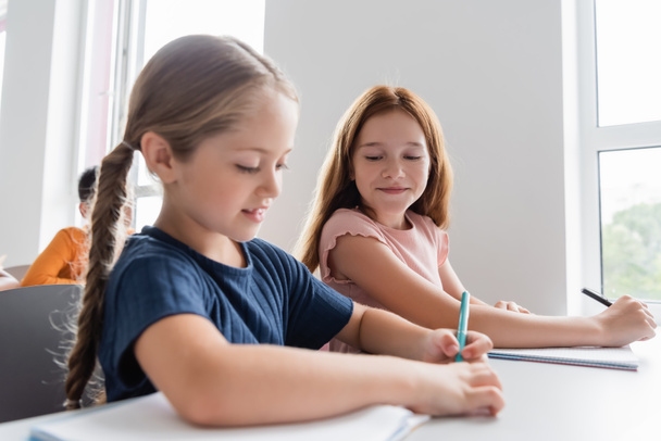 schoolgirl writing in notebook near smiling classmate during lesson - Foto, imagen