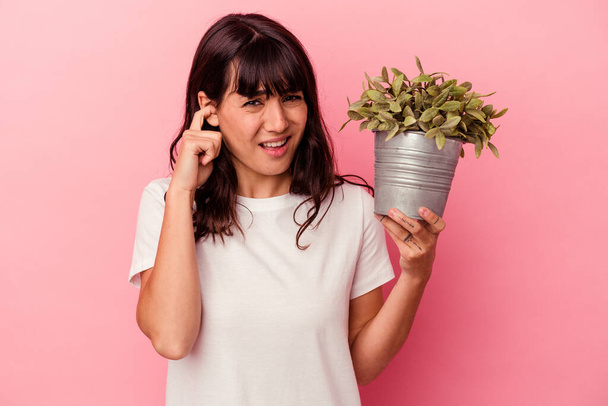 Young caucasian woman holding a plant isolated on pink background covering ears with hands. - Photo, Image