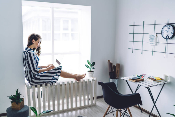 Side view of attractive young dark haired woman in striped dress sitting barefoot on wooden white windowsill and using laptop  - 写真・画像