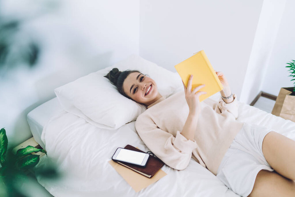 Glücklich lächelnde attraktive junge Frau in Homewear liegt im Bett neben dem Smartphone und schmiedet Pläne für den Tag, während sie Tagebuch checkt - Foto, Bild