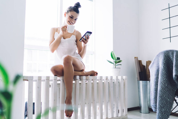 Content beautiful woman with hairbun sipping from mug and using smartphone while sitting at window sill in urban room with white walls in morning - Photo, Image