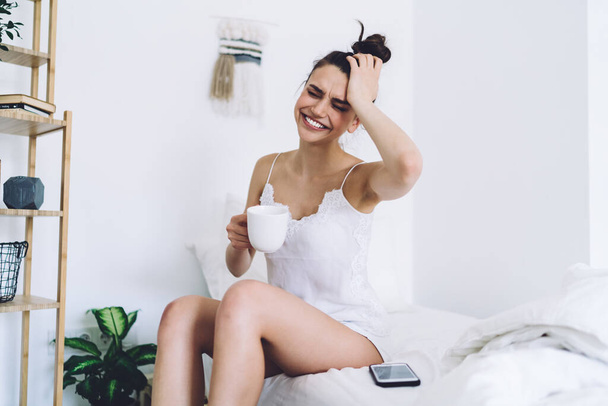 Frowning and young woman with hair bun in pyjamas carrying mug and rubbing head having migraine while sitting on bed in room with white walls - Photo, image