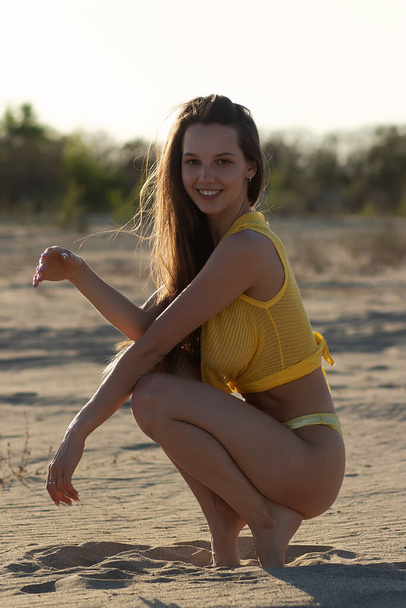 Pleine longueur de jeune voyageuse heureuse avec de longs cheveux foncés dans des maillots de bain jaunes élégants relaxant sur la plage de sable et regardant la caméra pendant les vacances d'été - Photo, image