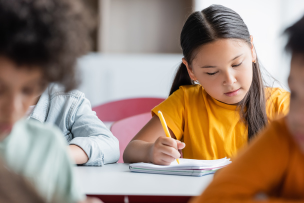 сфокусированная азиатская девушка пишет во время урока вблизи размытых мультиэтнических школьников - Фото, изображение