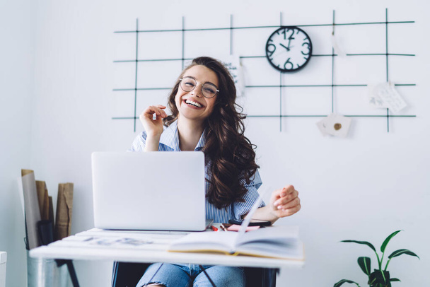 Cheerful adult dark haired woman in casual clothes and glasses looking at camera while sitting at desk and rising hand against white wall with black decor and clock in light minimalistic apartment - Valokuva, kuva
