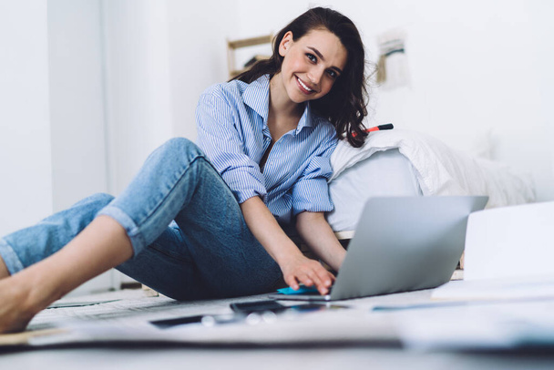 Side view of cheerful smiling woman in casual outfit typing laptop while sitting barefoot on floor with graphs and papers in modern room - Fotó, kép
