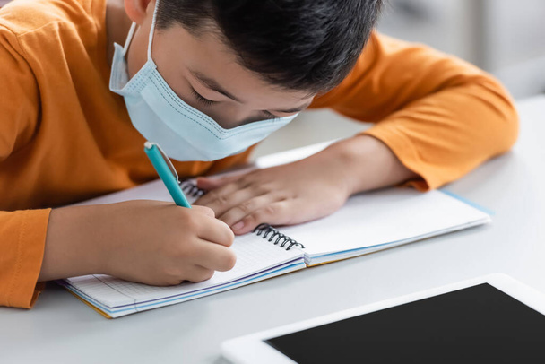 azjatycki chłopiec w masce medycznej pisanie w notatniku w pobliżu tabletki cyfrowej - Zdjęcie, obraz