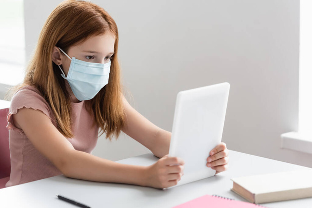 κορίτσι με ιατρική μάσκα κοιτάζοντας ψηφιακή ταμπλέτα κατά τη διάρκεια του μαθήματος σχολείο - Φωτογραφία, εικόνα