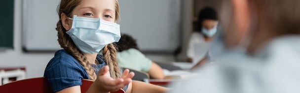 schoolgirl in medical mask pointing with hand near blurred schoolboy during lesson, banner - Фото, изображение