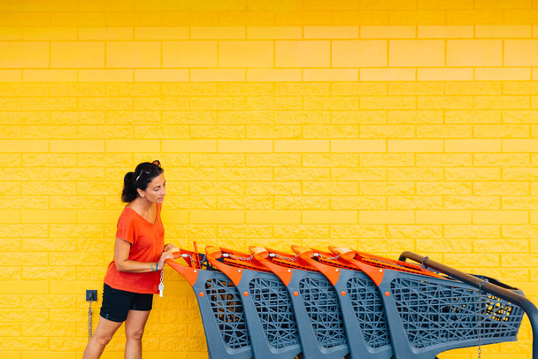 Женщина средних лет в красной одежде, берет тележку в супермаркете, на жёлтом фоне - Фото, изображение