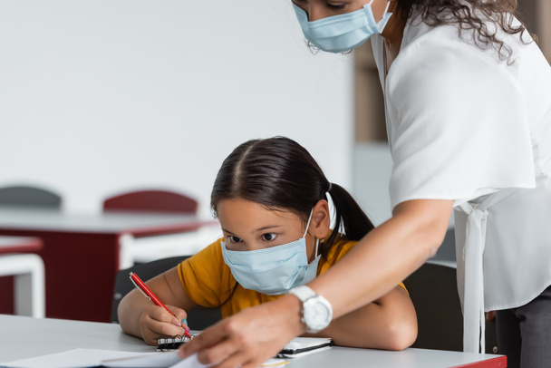 african american teacher in medical mask near asian girl writing in notebook - Zdjęcie, obraz