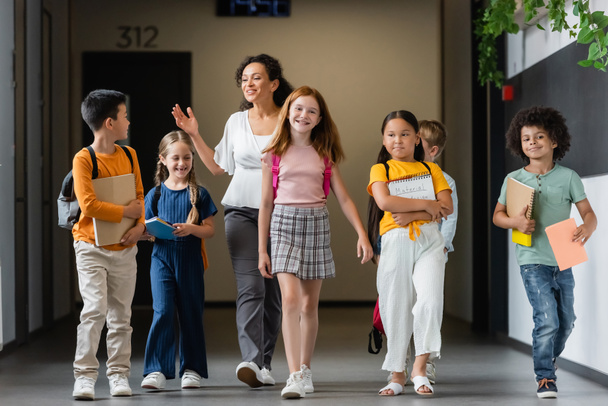 felici alunni multietnici che camminano lungo il corridoio scolastico con insegnante afro-americano - Foto, immagini