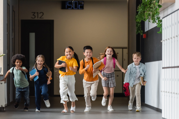 joyful multicultural friends with notebooks running in school corridor - Foto, afbeelding