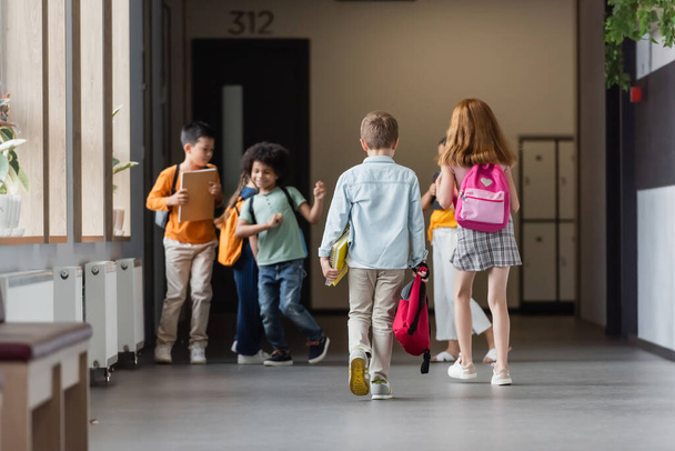 wieloetniczne dzieci w wieku szkolnym z plecakami i notatnikami w korytarzu szkolnym - Zdjęcie, obraz