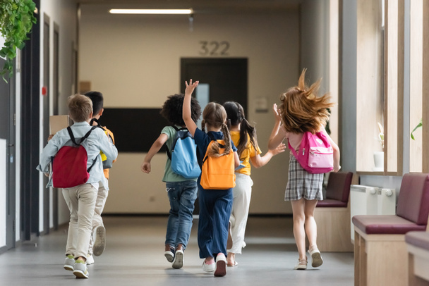 widok z tyłu wielonarodowych przyjaciół z plecakami biegającymi i machającymi rękami w korytarzu szkolnym - Zdjęcie, obraz