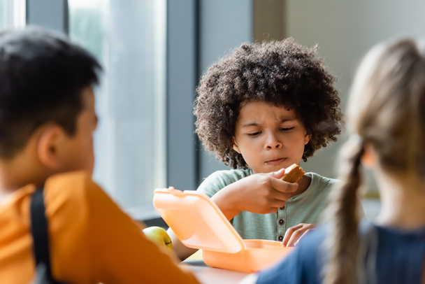 african american boy eating sandwiches near blurred classmates - Photo, Image