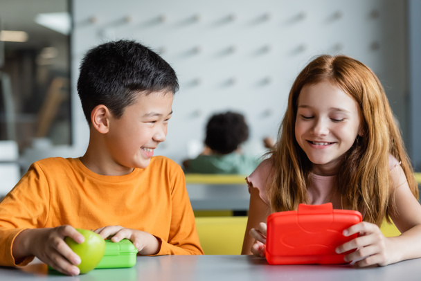 vörös hajú lány és mosolygós ázsiai fiú közel ebéd dobozok az iskolai étkezőben - Fotó, kép