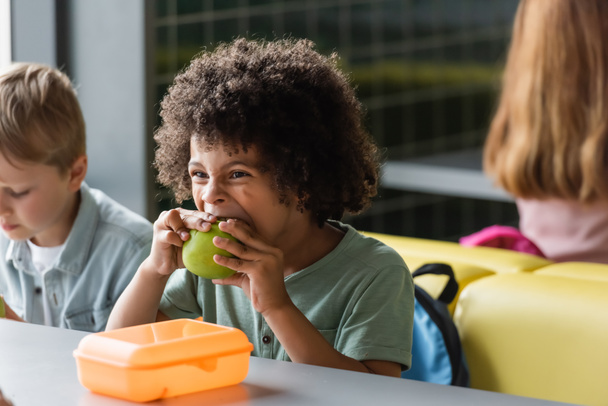 アフリカ系アメリカ人の少年が学校の食堂でぼやけたクラスメートの近くでリンゴを食べる - 写真・画像