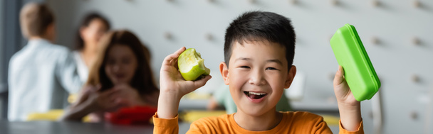 vrolijk aziatisch jongen holding lunch doos en appel in de buurt wazig kinderen in school eetgelegenheid, banner - Foto, afbeelding