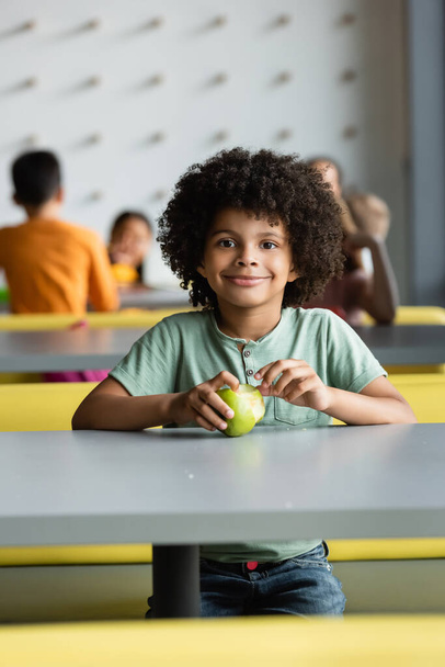african american schoolboy with fresh apple smiling at camera near blurred kids  - Photo, Image