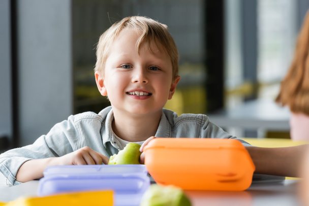 happy schoolboy looking at camera near lunch boxes on blurred foreground - Foto, Imagem
