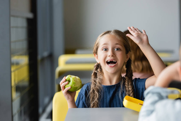 excited girl holding apple and waving hand in school eatery - Photo, Image