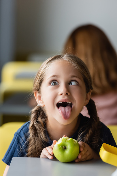 παιχνιδιάρικο κορίτσι με μήλο grimacing κατά τη διάρκεια του διαλείμματος γεύμα στο σχολείο τραπεζαρία - Φωτογραφία, εικόνα