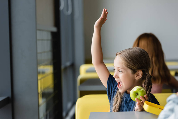 fille étonnée avec pomme fraîche agitant la main tout en regardant loin dans la cantine scolaire - Photo, image
