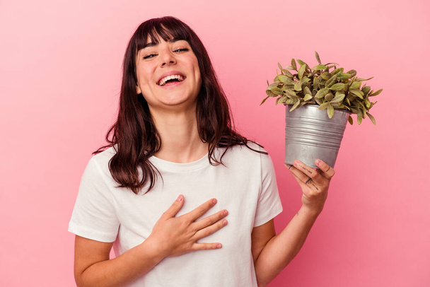 Молодая белая женщина, держащая растение на розовом фоне, громко смеется, держа руку на груди.. - Фото, изображение