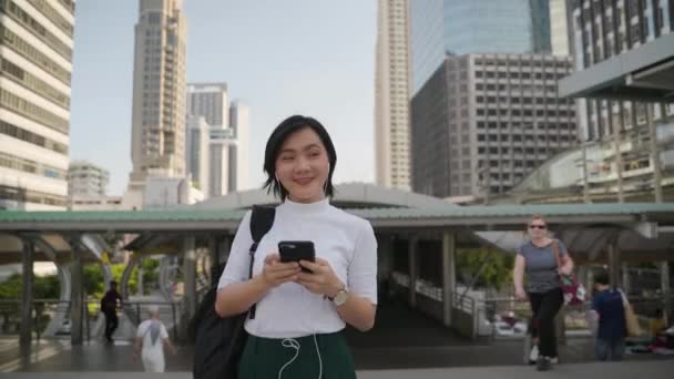 Portrait of asian woman with earphone listening music and using smartphone for chatting or browsing walks down the city street. Technology in everyday life and travel. - Footage, Video