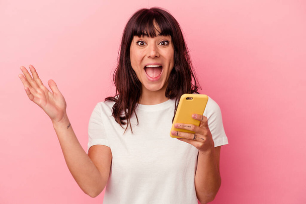Νεαρή καυκάσια γυναίκα που κρατά ένα κινητό τηλέφωνο απομονωμένο σε ροζ φόντο και δέχεται μια ευχάριστη έκπληξη, ενθουσιασμένη και σηκώνει τα χέρια. - Φωτογραφία, εικόνα