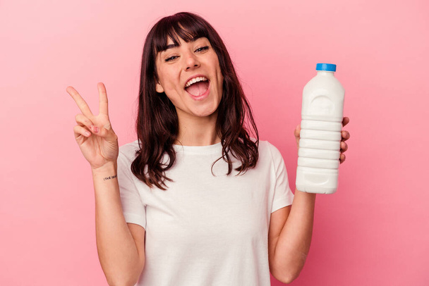 Joven mujer caucásica sosteniendo una botella de leche aislada sobre fondo rosa alegre y despreocupada mostrando un símbolo de paz con los dedos. - Foto, Imagen