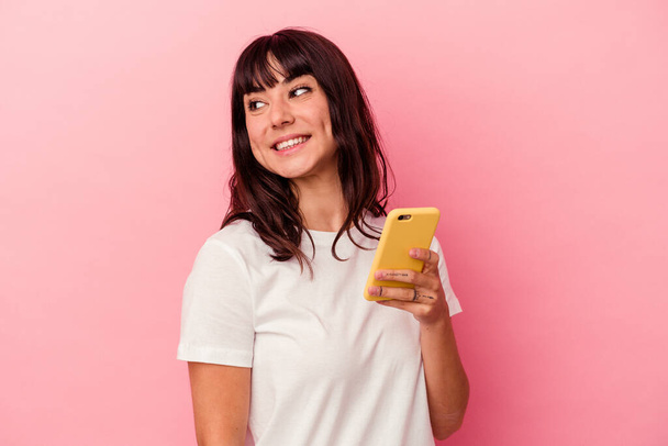 Joven mujer caucásica sosteniendo un teléfono móvil aislado sobre fondo rosa mira a un lado sonriente, alegre y agradable. - Foto, imagen