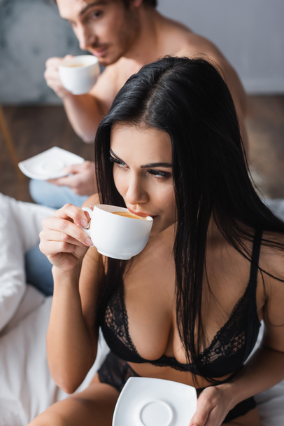 young woman in sexy bra drinking cappuccino near shirtless boyfriend on blurred background  - Photo, Image
