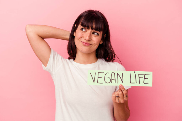 Joven mujer caucásica sosteniendo un cartel de vida vegana aislado en el fondo rosa tocando la parte posterior de la cabeza, pensando y haciendo una elección. - Foto, imagen