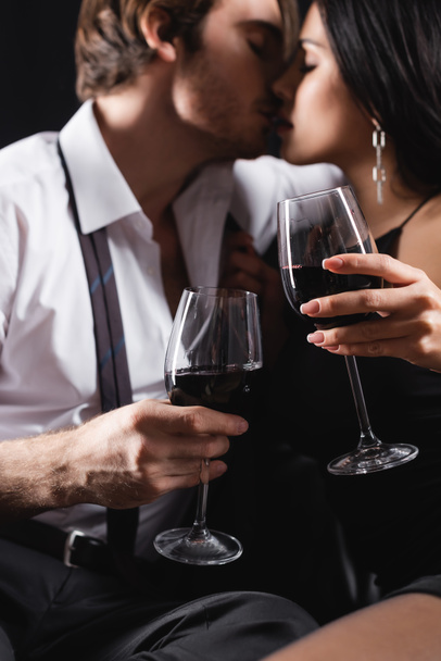 blurred and sexy couple holding glasses of red wine while kissing isolated on black  - Fotografie, Obrázek