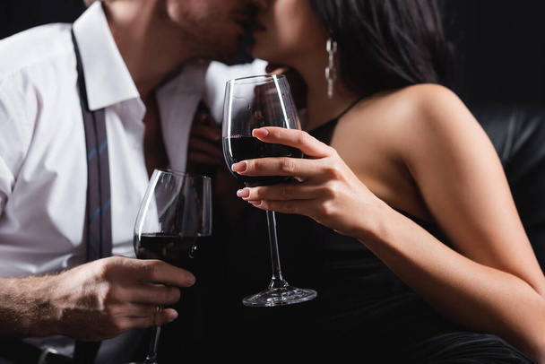 recortado vista de borrosa y sexy pareja sosteniendo vasos de vino tinto mientras se besa aislado en negro  - Foto, imagen