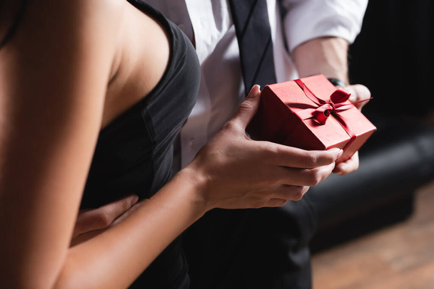 partial view of man holding red gift box near woman in dress on black - Foto, imagen