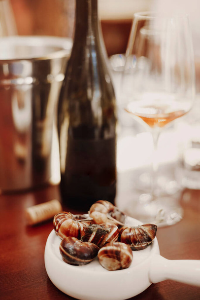 Pionowe ujęcie ślimaków podawane na białej ceramicznej patelni obok białego wina, smaczne ślimaki gotowane, tradycyjna francuska kuchnia w restauracji, wybiórczy nacisk na danie francuskie - Zdjęcie, obraz