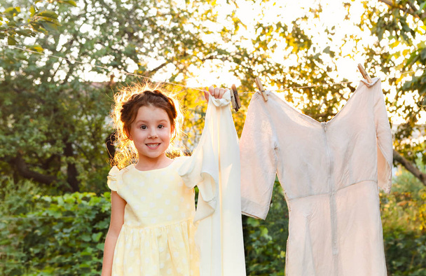 Petite fille mignonne en robe laver les vêtements blancs dans un bassin en métal dans la cour, aider les enfants avec les corvées, accrocher la lessive sur la corde à linge et le laisser sécher dans le jardin, sécher les vêtements à l'extérieur dans le vent - Photo, image