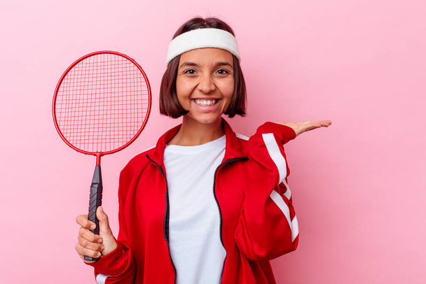 Junge Frau mit gemischter Rasse spielt Badminton isoliert auf rosa Hintergrund zeigt eine Kopie Raum auf einer Handfläche und hält eine andere Hand auf der Taille. - Foto, Bild