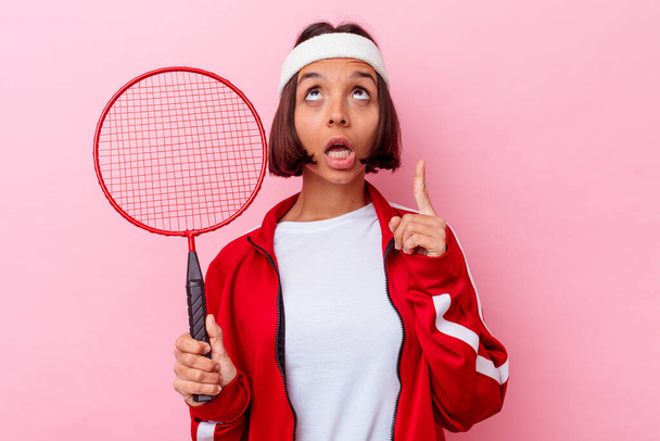 Mladý smíšený závod žena hraje badminton izolované na růžovém pozadí ukazuje vzhůru nohama s otevřenými ústy. - Fotografie, Obrázek