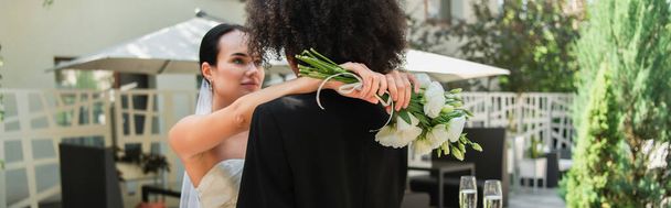 Young lesbian woman in dress holding bouquet while hugging african american girlfriend during wedding, banner  - Zdjęcie, obraz