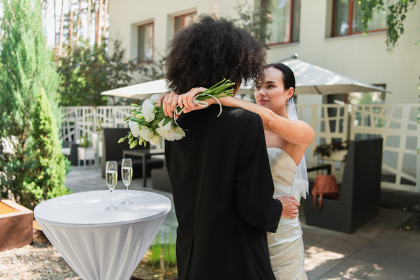 Lesbian woman holding wedding bouquet while hugging african american girlfriend in suit  - Foto, afbeelding