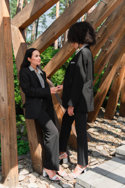 Lesbian woman in suit holding hand of african american girlfriend near arch in park  - Foto, Imagem